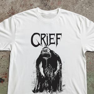 May include: A white short-sleeved t-shirt featuring a black graphic print on the chest. The print displays the word GRIEF in a stylised font above a dark, textured illustration of a creature with a wide, toothy mouth and a hunched posture.