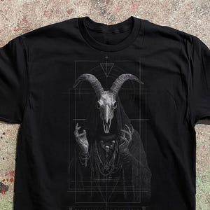 May include: A black t-shirt featuring a detailed white graphic print of a hooded figure with a goat skull head. The figure has raised hands and wears layered necklaces. The design is framed by thin white geometric lines, including triangles and rectangles.