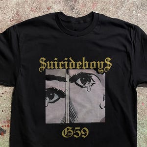 Op de afbeelding: Een zwart t-shirt met korte mouwen en een grafische print van twee ogen met een traan. De tekst $UICIDEBOY$ is in goud boven de afbeelding gedrukt en de tekst G59 is in goud onder de afbeelding op de voorkant van het shirt gedrukt.