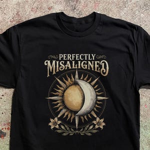 May include: A black short-sleeve t-shirt featuring a graphic print of a sun and crescent moon overlapping. The text "PERFECTLY MISALIGNED" is printed in a decorative font above the celestial design, which is flanked by two floral motifs.