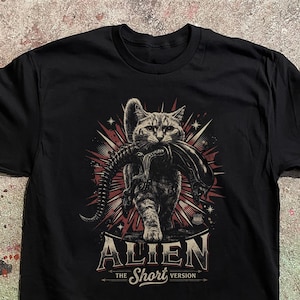 Pode incluir: Uma t-shirt preta com o gráfico de um gato malhado a carregar uma criatura alienígena na boca. O design inclui um fundo de explosão estelar vermelho e branco, o texto ALIEN em letras grandes e o texto THE SHORT VERSION por baixo.