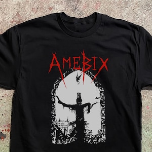Puede incluir: Una camiseta negra de manga corta con un estampado gráfico en rojo, blanco y negro. El texto rojo dice AMEBIX sobre un marco de ventana arqueado blanco que contiene una silueta negra encadenada con el texto WE ENY NO MASTER.