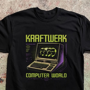 Puede incluir: Una camiseta negra de manga corta con un estampado gráfico de un monitor de ordenador antiguo que muestra cuatro rostros. El texto KRAFTWERK y COMPUTER WORLD aparece en verde lima. Incluye MAN MACHINE MUSIC y detalles de rejilla técnica.