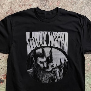 Puede incluir: Una camiseta negra de manga corta con un estampado gráfico en tonos grises. El diseño incluye el texto "ELECTRIC WIZARD" en una fuente estilizada sobre una ilustración circular de una figura barbuda y con cuernos que sostiene un cáliz humeante.