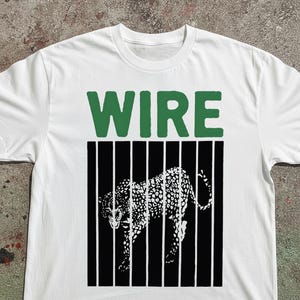 May include: A white short-sleeve t-shirt featuring a graphic print on the chest. The design includes the word WIRE in large green capital letters above a black and white illustration of a spotted leopard standing behind vertical black bars.