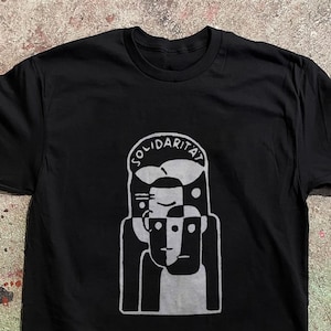 以下が含まれることがあります： 胸元にグレーのグラフィックプリントが施された黒の半袖Tシャツ。デザインは、丸みを帯びたフレームの中に抽象的で様式化された人の顔が縦に積み重なっており、グラフィックの上部にはアーチ状にSOLIDARITÄTという文字がプリントされています。
