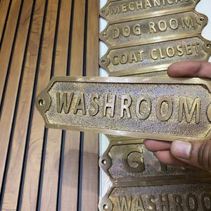 May include: A rectangular brass sign with a textured finish and raised lettering that reads WASHROOM. The sign features rounded ends with small holes for mounting. Several similar brass signs with various labels are visible in the background.