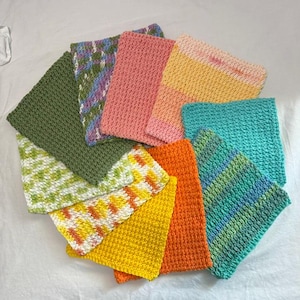 May include: A collection of ten rectangular crochet washcloths arranged in a fan shape. The cloths feature various solid colors including olive green, pink, orange, and yellow, alongside variegated patterns in shades of blue, green, and white.