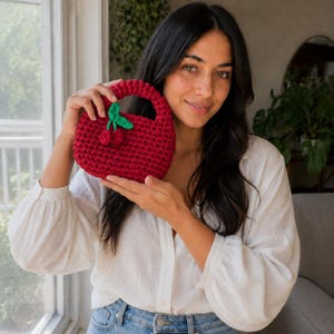 Pode incluir: Uma pequena bolsa de mão vermelho-escura, feita à mão em crochê, com uma alça arredondada e um par decorativo de pequenas cerejas vermelhas texturizadas com um caule verde brilhante e duas folhas presas à superfície externa da bolsa.