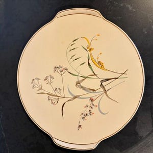 Op de afbeelding: Een ronde, crèmekleurige keramische serveerschaal met twee geïntegreerde handgrepen. Het oppervlak is voorzien van een delicate botanische illustratie van wilde grassen en bloemen in bruin, geel, groen en grijs, met een dunne bruine rand.