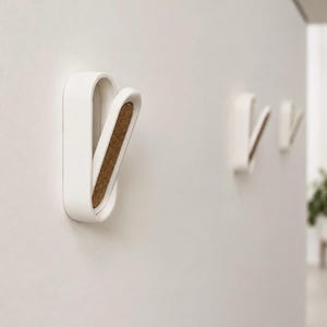 Peut inclure: Trois patères murales blanches de forme ovale allongée et arrondie. Chaque patère est dotée d'un bras articulé à ressort qui s'ouvre vers l'extérieur, révélant une incrustation en liège brun texturé sur la surface intérieure du mécanisme.