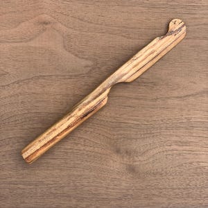 May include: A hand-carved wooden spreader with a natural light brown finish and visible dark wood grain patterns. The utensil features a cylindrical handle that tapers into a flat, slightly curved blade with a rounded tip for spreading.