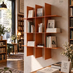Op de afbeelding: Een aan de muur gemonteerde oranje metalen rasterplank met verticale en horizontale verdelers. De planken bevatten diverse boeken, waaronder titels als The Daily Stoic en At Home in the World, samen met een kleine houder en een bril.