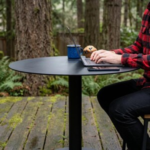 Pode incluir: Uma mesa redonda de pedestal em preto mate com um tampo fino e uma base cilíndrica. A superfície da mesa contém um computador portátil prateado, uma caneca de esmalte azul salpicado com vapor, um muffin de mirtilo e um smartphone preto.