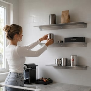 Op de afbeelding: Drie horizontale wandplanken van geborsteld metaal gemonteerd aan een wit betegelde muur. De planken bevatten diverse keukenartikelen, waaronder een bruine papieren zak, een zwarte Nespresso-doos, zilveren bussen en twee zilveren metalen mokken.