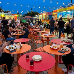 May include: An outdoor dining area featuring multiple round orange and red tables with matching metal chairs. Several food trucks are parked in the background under strings of glowing lights. People are seated at the tables eating tacos and chips.