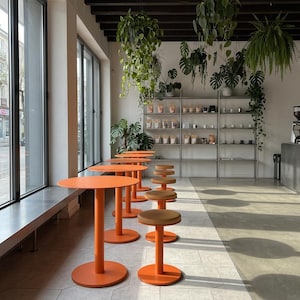 May include: A series of bright orange circular tables and matching stools arranged in a row within a spacious room. The space features large windows, light-coloured tiled flooring, and metal shelving units filled with various products.