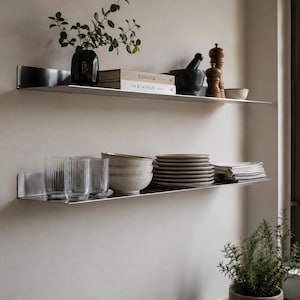 Puede incluir: Dos estantes de pared horizontales de metal cepillado montados uno sobre otro. El estante superior contiene un jarrón negro con vegetación, dos libros titulados KINFOLK, un mortero negro con mano y molinillos de madera. El estante inferior contiene vasos estriados, cuencos, platos y servilletas.
