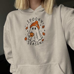 Könnte beinhalten: Ein beigefarbener Kapuzenpullover mit einer Grafik eines Geistes, der eine orangefarbene Strickmütze trägt und eine Tasse mit Kürbisgesicht hält. Der Text SPOOKY SEASON ist in Schwarz um den Geist herum gedruckt, umgeben von kleinen Herbstblättern.