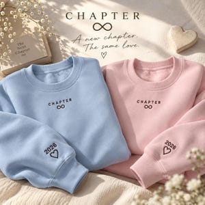 Op de afbeelding: Een lichtblauw en een lichtroze sweatshirt, beide voorzien van donkerbruin borduursel op de borst met de tekst "CHAPTER" boven een oneindigheidssymbool. Op de mouw van elk sweatshirt staat het jaartal "2026" boven een hartvorm.