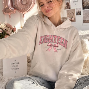 May include: A beige hooded sweatshirt featuring the text EIGHTEEN in pink letters with a pink bow graphic underneath, accompanied by the text EST. 2008. The hoodie has long sleeves, a front kangaroo pocket, and a drawstring hood, made of cotton fabric.