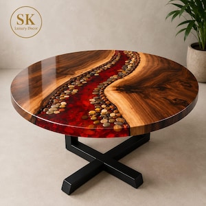 May include: A round coffee table featuring a polished wood surface with a central river-like inlay of red resin embedded with smooth, multi-coloured river stones. The table is supported by a black metal base with a cross-shaped design.