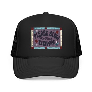 May include: A black trucker hat featuring a rectangular graphic on the front panel. The graphic has a purple background with the text "PLEASE SLOW THE FUCK DOWN" in a dark, wavy font, surrounded by a decorative border with small floral accents.