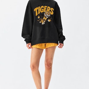 Puede incluir: Una sudadera negra de manga larga con cuello alto y un estampado gráfico de un tigre con un suéter con la letra M, acompañado del texto MIZZOU TIGERS en amarillo. Combinado con pantalones cortos amarillos con un patrón de lunares blancos.