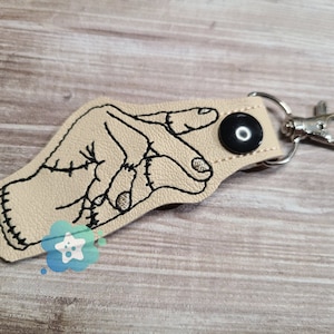 May include: Beige leather keyring with a black embroidered hand making a peace sign. The keyring has a silver metal ring.