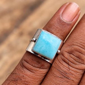 May include: A wide silver ring featuring a square, polished light blue Larimar gemstone set in a smooth metal bezel. The band has a flat, polished surface with small decorative metal spheres on either side of the stone setting.