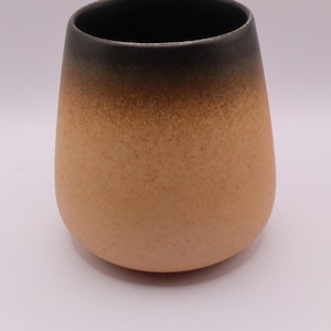 May include: A rounded ceramic cup featuring a speckled texture with a color gradient that transitions from a light tan base to a dark brown rim. The interior of the cup is dark brown, and the exterior surface displays a matte finish with visible dark speckles throughout the tan and brown sections.
