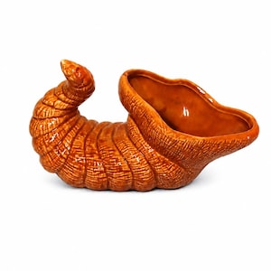 May include: A glossy, orange-brown ceramic cornucopia vessel featuring a textured, ridged exterior surface that mimics the appearance of woven material. The horn-shaped container has a wide, open mouth and a curved, tapered end.