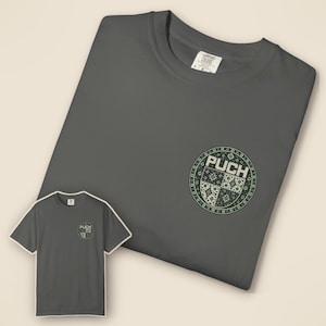 Könnte beinhalten: Ein anthrazitfarbenes Kurzarm-T-Shirt mit einer kreisförmigen Grafik auf der linken Brust. Die Grafik zeigt den Text PUCH in weißen Blockbuchstaben über einem viergeteilten Muster aus dunkelgrünen und weißen geometrischen Formen und Blumen.