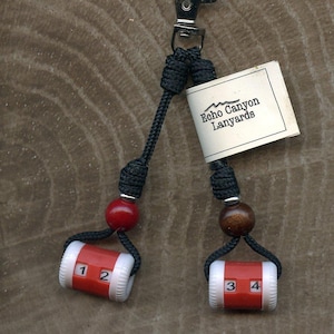 May include: A detachable double flush counter featuring two black cords attached to a metal lobster clasp. Each cord has a wooden bead and a red and white cylindrical counter displaying the numbers 1, 2, 3, and 4. A white tag reads Echo Canyon Lanyards.