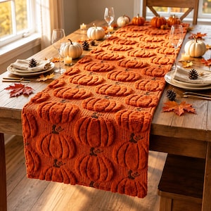 May include: A long, rectangular table runner in a vibrant orange colour, featuring a repeating, raised, textured pumpkin pattern across its entire surface. The runner is made of a knit fabric and is draped over a wooden dining table.