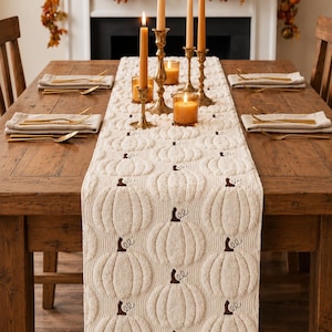May include: A cream-coloured quilted table runner featuring a repeating embossed pumpkin pattern with dark brown embroidered stems. The runner is laid flat on a wooden dining table, centred between place settings with gold-toned cutlery.