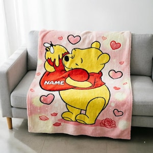 May include: A soft, light pink fleece blanket featuring a large yellow bear character wearing a red shirt, holding a honey pot with the word NAME printed on it. The design includes scattered pink hearts and small rose illustrations throughout.