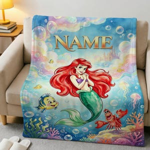 Puede incluir: Una manta de forro polar suave con una colorida escena submarina que muestra una sirena de pelo rojo, un pez amarillo y azul, y un cangrejo rojo. El diseño incluye burbujas flotantes, coral, un castillo y la palabra NAME en letras doradas.
