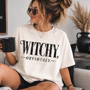 May include: An off-white short-sleeve t-shirt featuring the black text "WITCHY, OBVIOUSLY." printed in a distressed serif font across the chest. The garment has a crew neckline and a relaxed fit, constructed from a solid-colored fabric.
