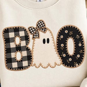 Puede incluir: Una sudadera color crema con un diseño de aplique bordado que deletrea BOO. La letra B es de cuadros negros y blancos, la letra O es negra con un patrón floral blanco y el fantasma central tiene un lazo de cuadros.