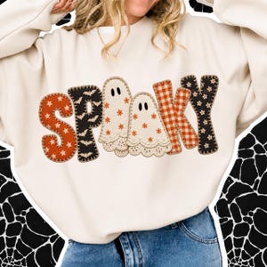 May include: A cream-coloured long-sleeve sweatshirt featuring the word SPOOKY across the chest. The letters are designed with various patterns including orange stars, black bats, cream ghosts with orange flowers, and an orange and white gingham check.
