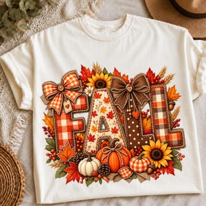 Op de afbeelding: Een crèmekleurig t-shirt met het woord FALL in grote, decoratieve letters. De letters zijn versierd met ruitpatronen, herfstbladeren, zonnebloemen, pompoenen, dennenappels en strikken in oranje, bruine en gele tinten.
