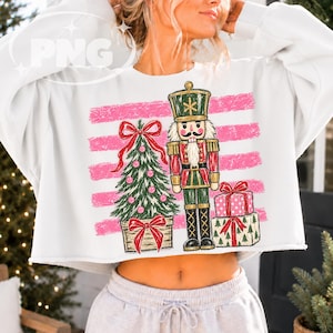 May include: A white long-sleeve cropped sweatshirt featuring a graphic print of a nutcracker, a decorated Christmas tree, and two stacked gift boxes set against a background of horizontal pink distressed stripes.