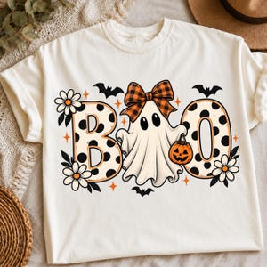 Könnte beinhalten: Ein cremefarbenes T-Shirt mit einer Grafik und dem Schriftzug BOO. Die Buchstaben B und O sind cremefarben mit schwarzen Punkten. Ein weißer Geist mit einer orange-schwarzen karierten Schleife trägt einen Kürbiseimer. Schwarze Fledermäuse, weiße Blumen.