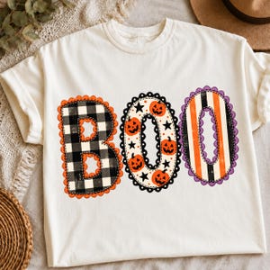 May include: A cream-colored short-sleeve t-shirt featuring the word BOO in large, decorative letters. The B is black and white buffalo plaid with an orange scalloped border, the O is white with orange pumpkins and black stars, and the final O is orange and black stripes with a purple scalloped border.