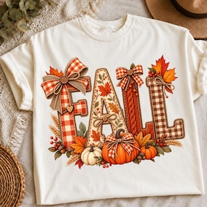 Op de afbeelding: Een crèmekleurig t-shirt met het woord FALL in grote letters. De letters zijn versierd met rood-witte ruitpatronen, herfstbladeren, tarwe, eikels en oranje pompoenen. Een grote ruitjesstrik zit boven op de letter F.