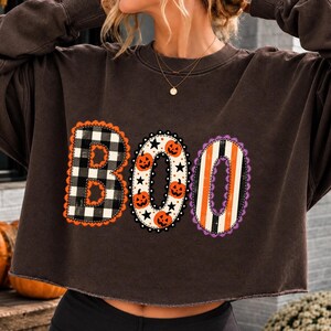 Puede incluir: Una sudadera de manga larga de color marrón oscuro con la palabra BOO impresa en la parte delantera. La letra B tiene un patrón de cuadros blanco y negro, la letra O muestra calabazas naranjas y estrellas negras, y la última O tiene rayas naranjas y negras.