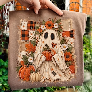 May include: A brown sweatshirt featuring a central embroidered design of a ghost holding a pumpkin. The ghost is surrounded by sunflowers, daisies, autumn leaves, and a patchwork background of orange and black plaid fabric squares.