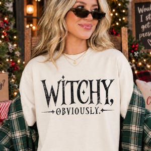 May include: A cream-colored crewneck t-shirt featuring the black distressed text WITCHY, OBVIOUSLY. The design includes small star accents above and below the words. The shirt is worn with a green and black plaid flannel shirt draped over the shoulders.