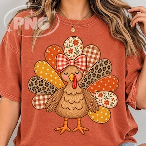 May include: A rust-colored short-sleeve t-shirt featuring a central graphic of a turkey with closed eyes and a red and white checkered bow. The turkey's tail feathers display various patterns including leopard print, polka dots, gingham, and floral.